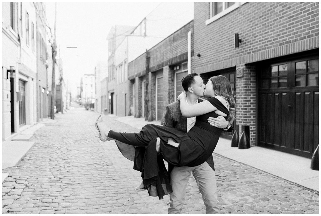 A man lifts a woman up an d kisses her on a cobblestone street.