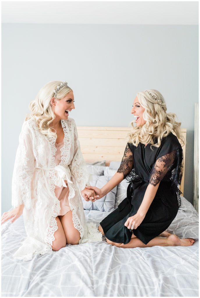 The bride and her twin sister maid of honor hold hands bouncing on the bed with excitement on the wedding day morning.