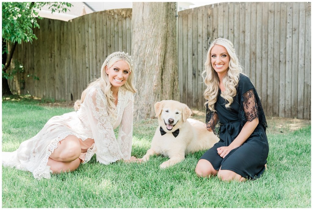 The bride and her maid of honor sit with their dog in the backyard while wearing robes.