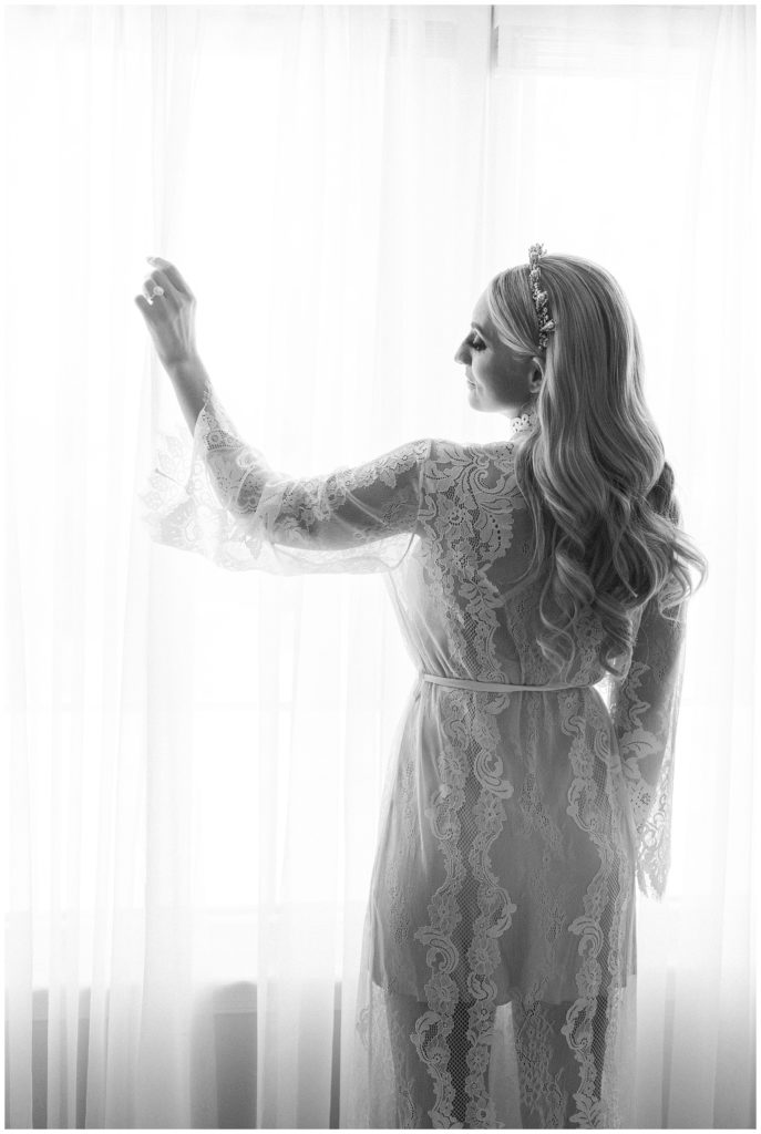 The bride pulls back a curtain in her bedroom while wearing her lacy wedding day robe.