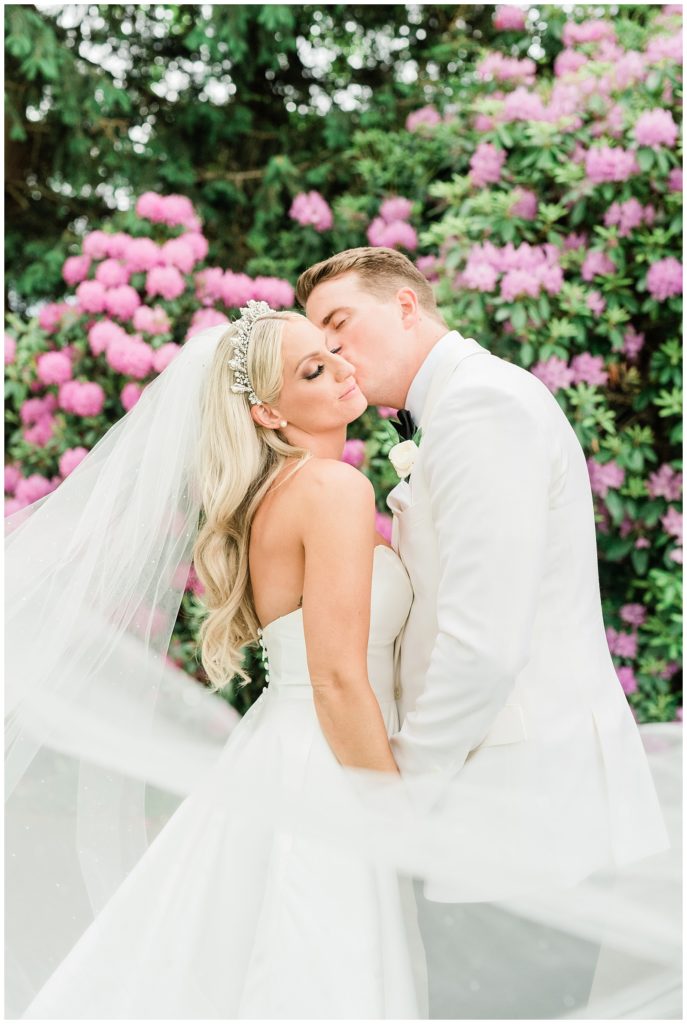 Groom kisses the bride on her cheek in front of a floral bush while she wears her Maria Elena bridal headpiece.