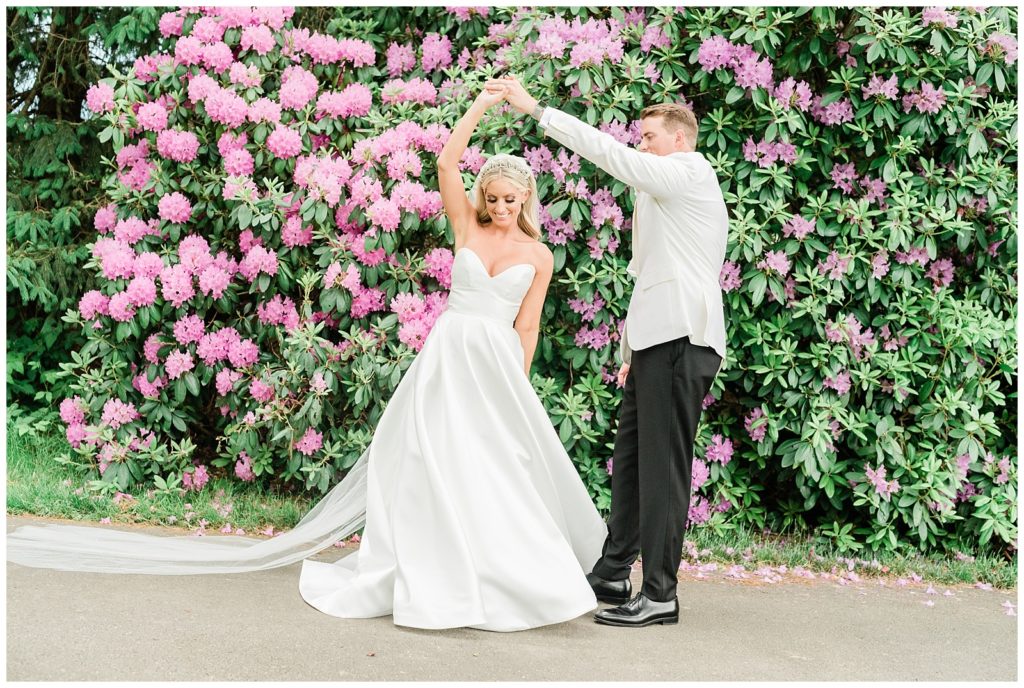 The groom twirls the bride in front of a rhododendron bush.