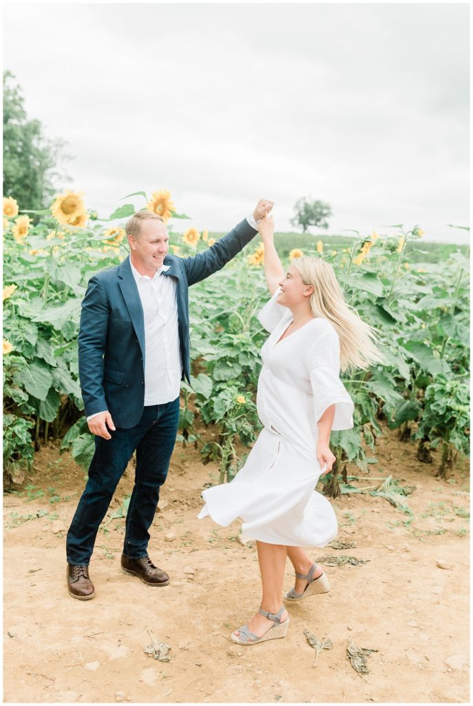 Rachel and Brian are on a dirt path, with sunflowers behind them. Brian is holding Rachel's arm up and she appears to be playfully dancing. Her hair flows behind her as she looks twoards him. 