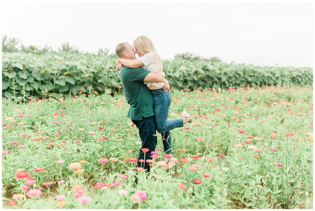 Surrounded by pink sunflowers, Brian is holding Rachel. She has her arms around his neck and he is holding her up by the waist with her one leg kicking backwards. 
