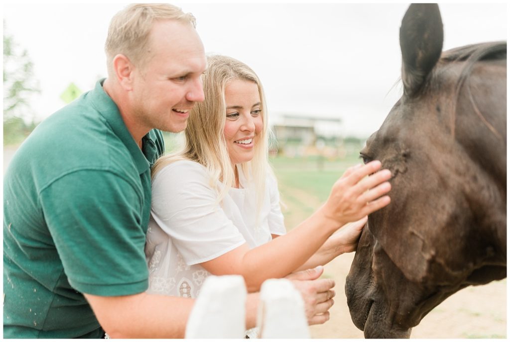 Brian is standing behind Rachel as she reaches forward with her right arm to touch the side of a horse's face. She is focused on the horse and smiling. Brian reaches around her and holds his hand just below the horses nose. 