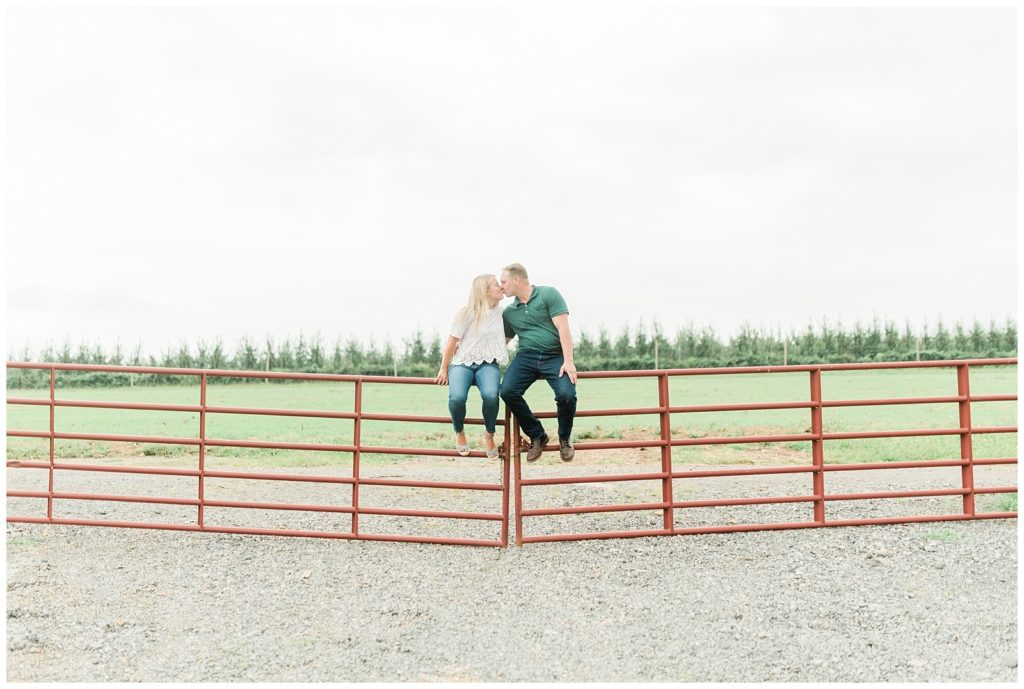 Rachel and Brian are sitting on the corner of a metal barn style fencing intended for horses. They lean in and kiss each other. There is a vibrant green tree line in the far background and a grass field behind the fence. 