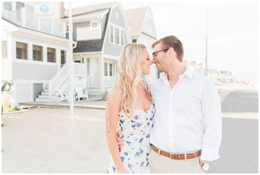 Kristin and Ryan stand slightly turned towards each other with their arms around each other, faces pressed nose to nose and smiling. They stand in front of blue beach homes with a light sky.