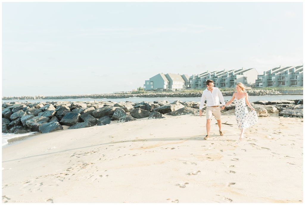 Ryan and Kristin walk hand in hand along the sand of the Manasquan Beach. Rocks lining the ocean are visible on the left side of the image. The ocean is visible in the background with shore homes lining the water edge. 