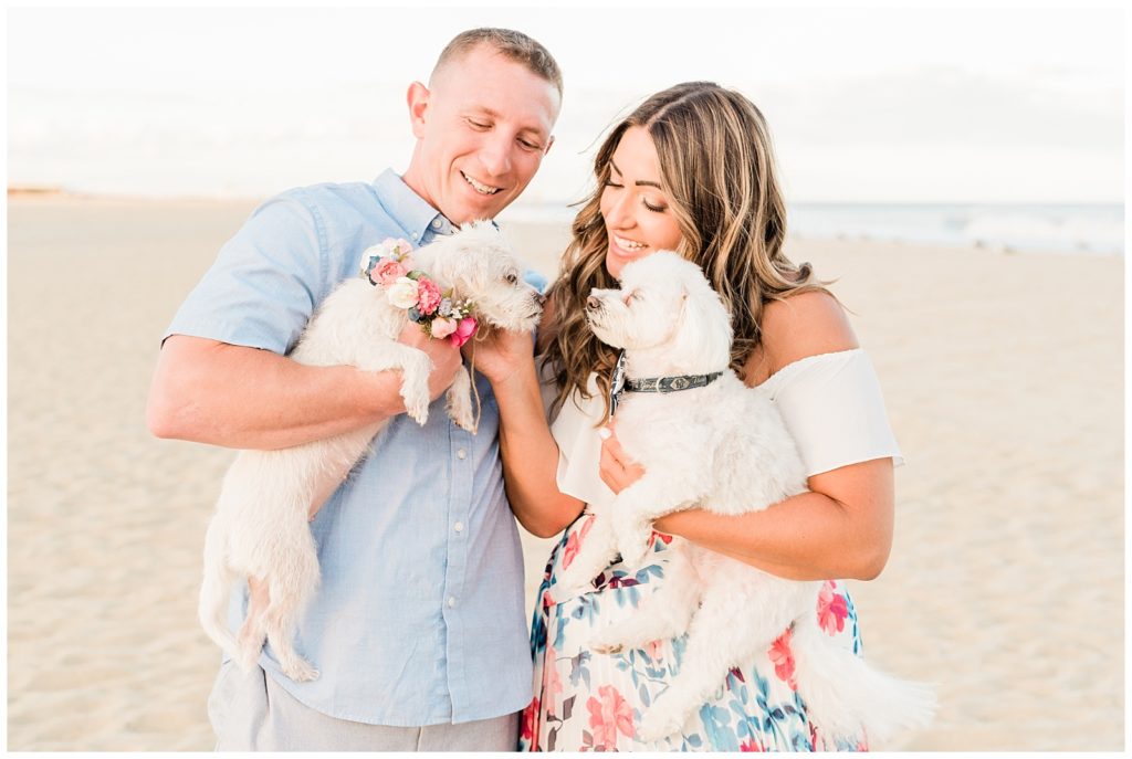 Jim and Alessandra stand on the beach each holding a pup. They both are looking down at the pup in Jim's arms fondly while the pup is wiggling up towards their faces for kisses. 