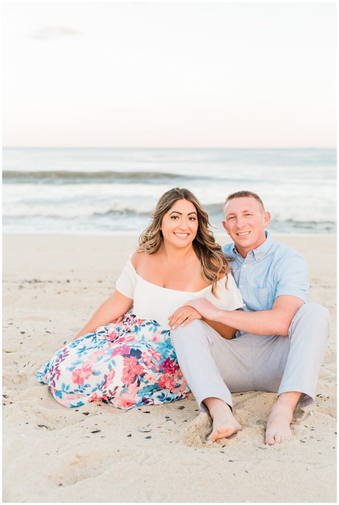 Alessandra and Jim sit in the sand. Jim's arm is behind her, and her hand is on his right leg. His other hand covers the top of hers as they both face forward. The ocean waves crash on the shore behind them. 