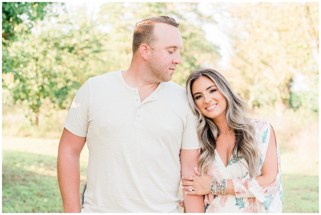 Nicolette and Mike are standing side by side. She rests her head on his shoulder, and her arms are loosely wrapped around his. She is facing forward smiling while he looks down towards the top of her head. 