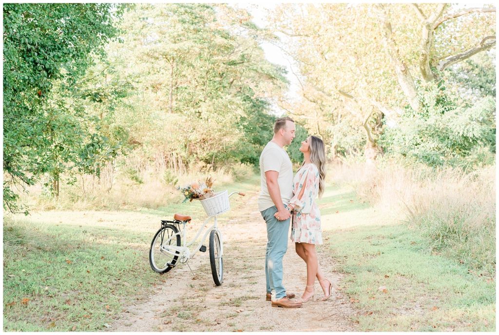 Nicolette and Mike stand close, facing each other hand in hand on a grassy pathway with worn edges. The bicycle is placed just off to the left. 