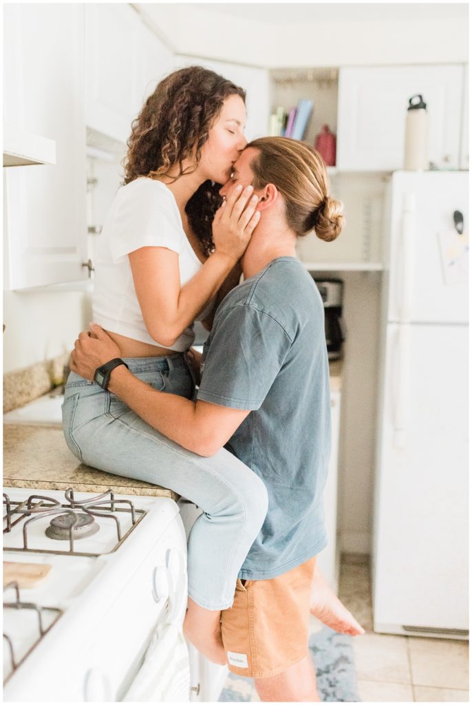 Thiana sits on teh kitchen counter top. Brandon has his arms around her waist. Her hands cup the side of his face as she kisses his forehead. 