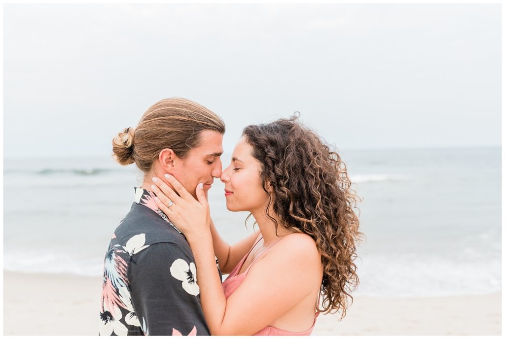 Thiana and Brandon on the beach with teh ocean visible behind them. This side angled shot shows them nose to nose, with Thiana's hands cupping the sides of his face. 