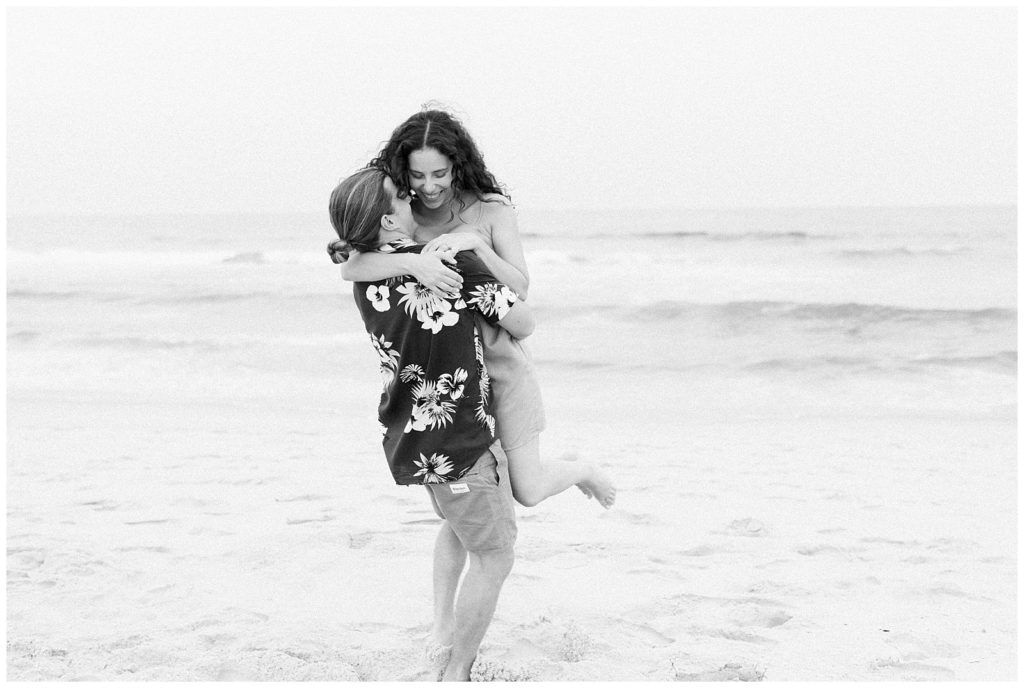 A balck and white image of Brandon standing on the beach with tThiana in his arms. He has her lifted in the air and her legs curled behind her. 