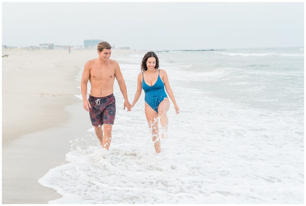 Thiana and Brandon in bathing suits walking quickly hand in hand through the breaking ocean waves along the shoreline. 
