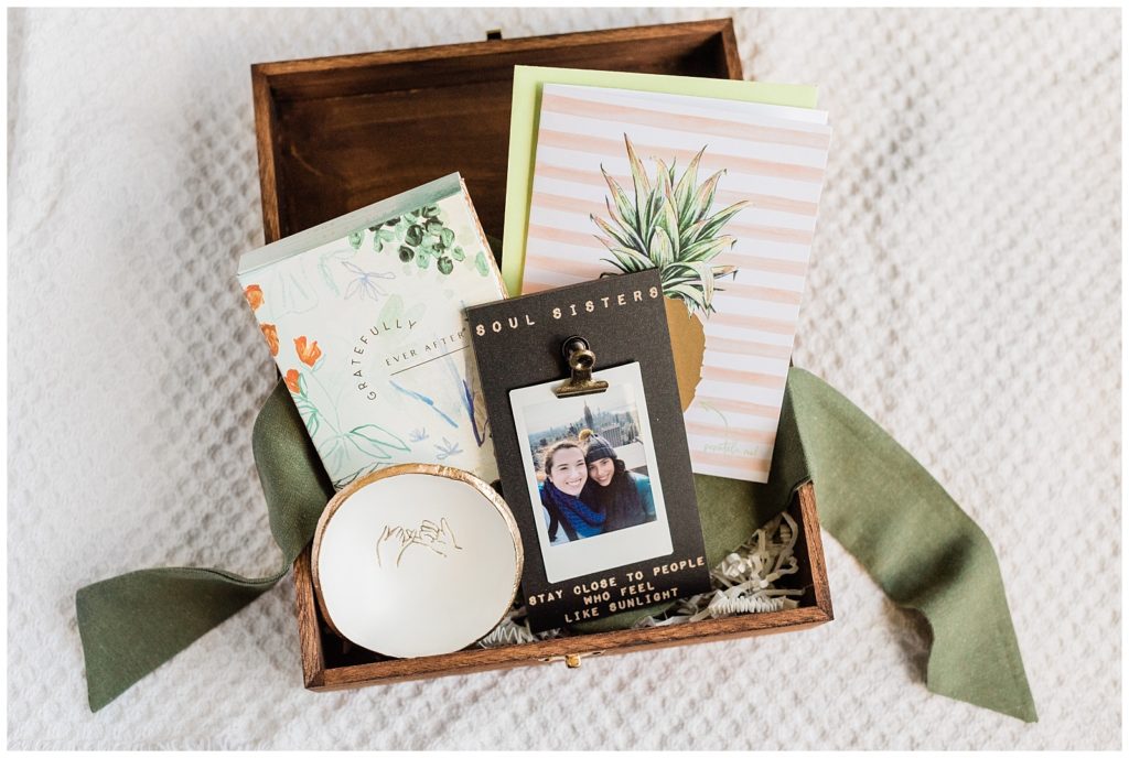 A Polaroid print, a jewelry dish, perfume, ribbon and pineapple scratch off card are laid out for a bridesmaid proposal gift.