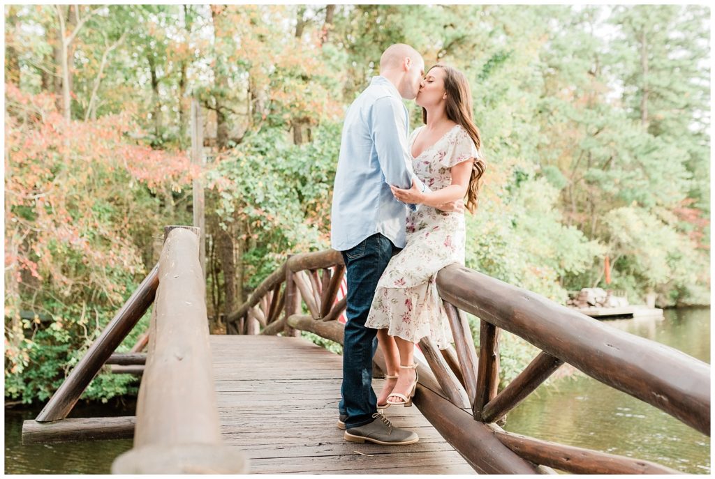 Kellie and Michael are standing on a wooden bridge. She is leaning against the railing and him against her as they closely hug. 