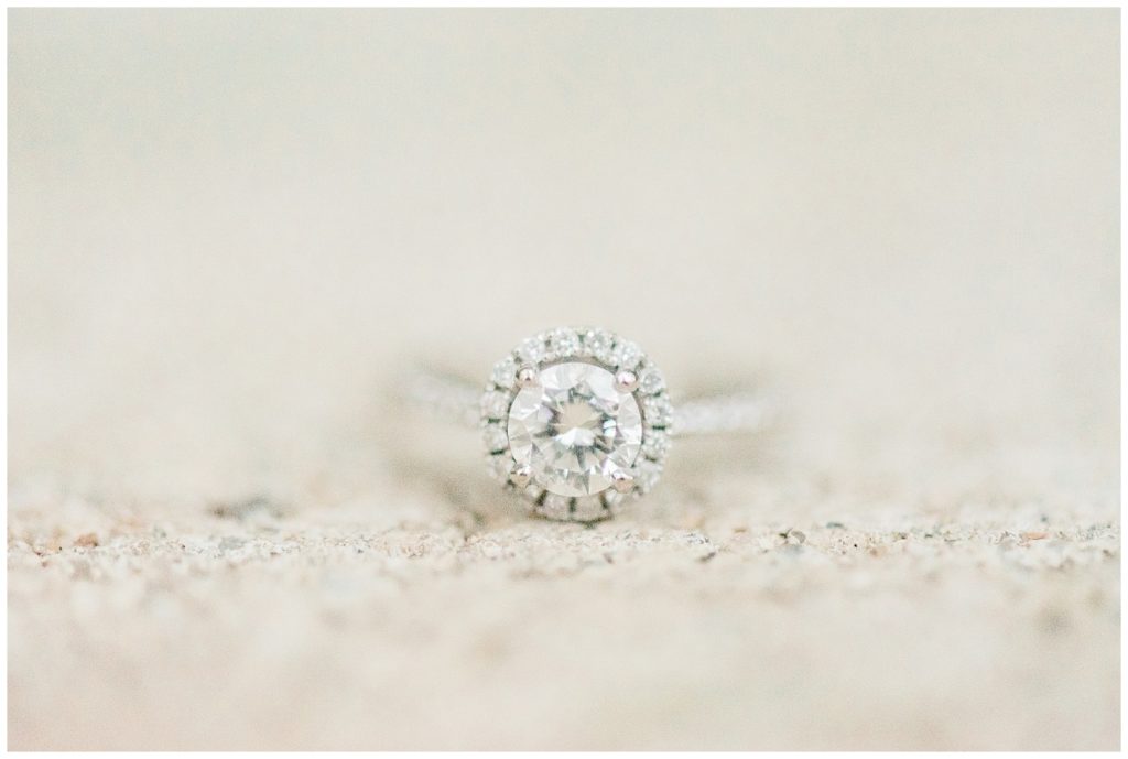 A close up shot of a circular engagement ring. The setting has small diamonds around the outside and a central, pronounced diamond in the center. 