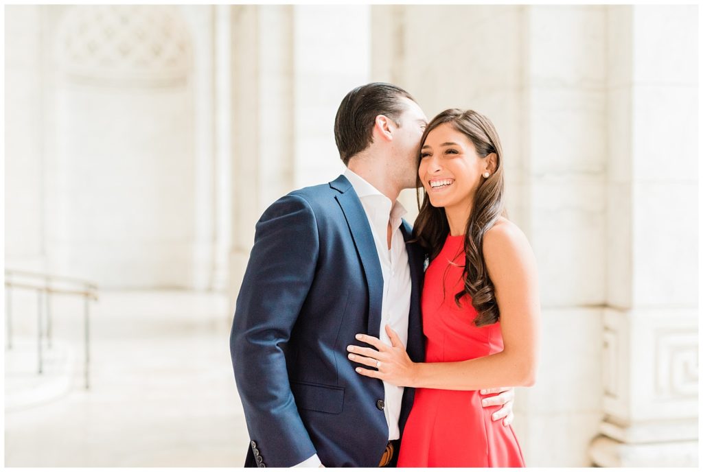 Nicole and Mike stand in front of a white pillar of the NY public library. Nicole has her hand on Mike's chest as he leans in to kiss her cheek