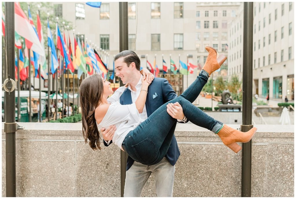 In front of a fountain on the streets of NYC, Mike is holding Nicole in his arms. Her legs are wrapped in his left hand and her upper body in his right. Her legs are kicked up just a bit as he leans down a little closer to her face as they stare into each other's eyes. Her arms are wrapped around his neck. 