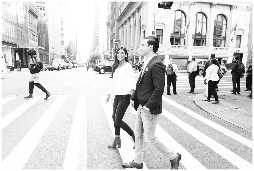 A black and white image of Nicole and Mike walking on a crosswalk in an NYC street. Mike is walking straight ahead and Nicole is looking at him as the image is captured mid stride. 