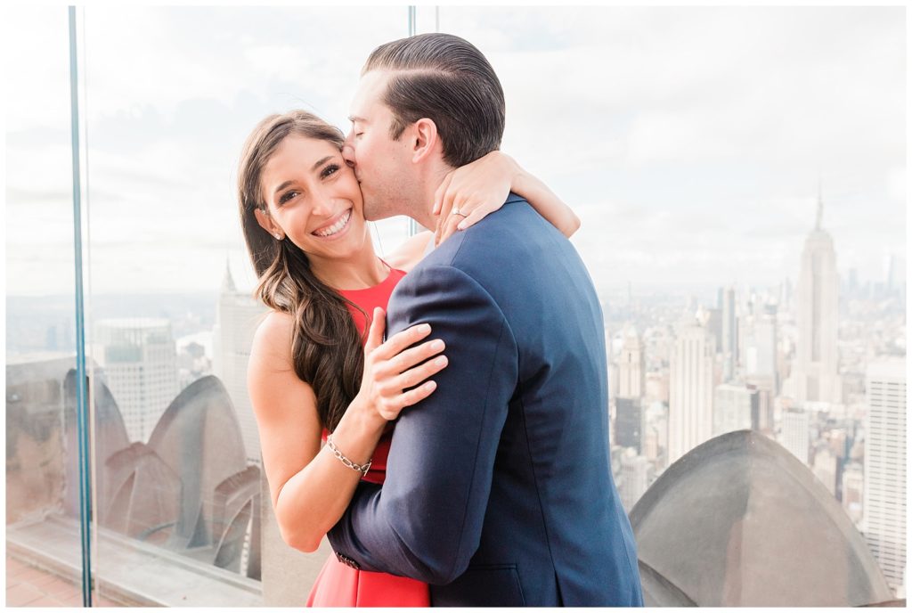 Nicole's back is against the wall at top of the rock. You can vaguely see the city skyline in the background. She is smiling and facing the camera, with her hands on Mike's shoulders. Mike is standing in front of her, kissing her temple with his arms around her waist. 