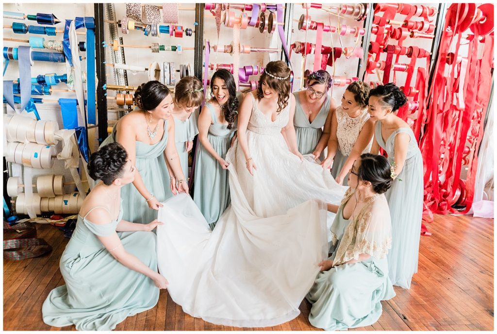 Jillian surrounded by her bridesmaids either standing or kneeling and holding the hems of her long and flowing dress. 