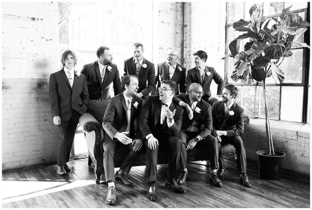 a black and white image of the groom and all groomsmen. Several are standing in one row and a more forward row is sitting on a couch. All are dressed in suits. 