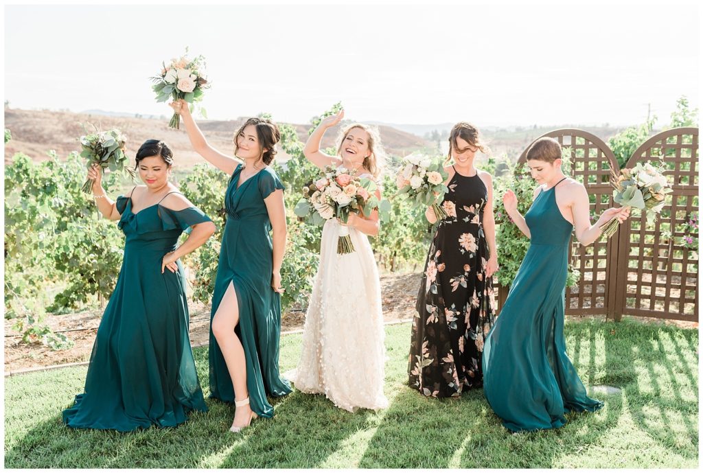 A bride celebrates with her bridesmaids wearing teal dresses, and her maid of honor wearing a black floral print dress outside at Avensole Winery in Temecula, CA.
