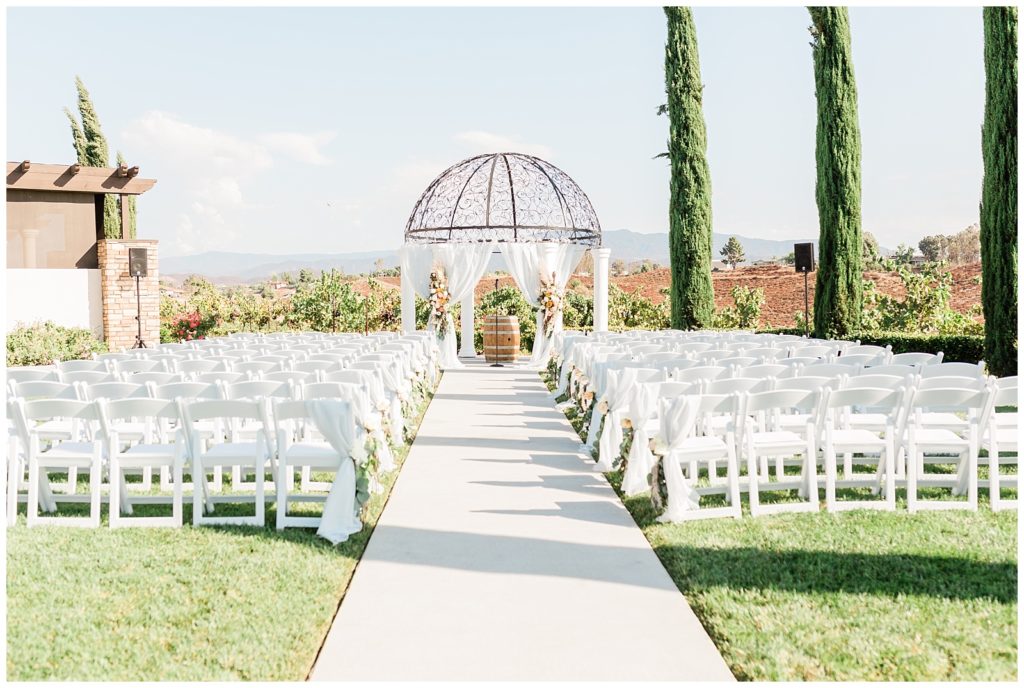 Outdoor ceremony space is set for a wedding with a wine barrel under the main arch and white folding chairs for guests, at Avensole Winery wedding venue in Temecula, California.