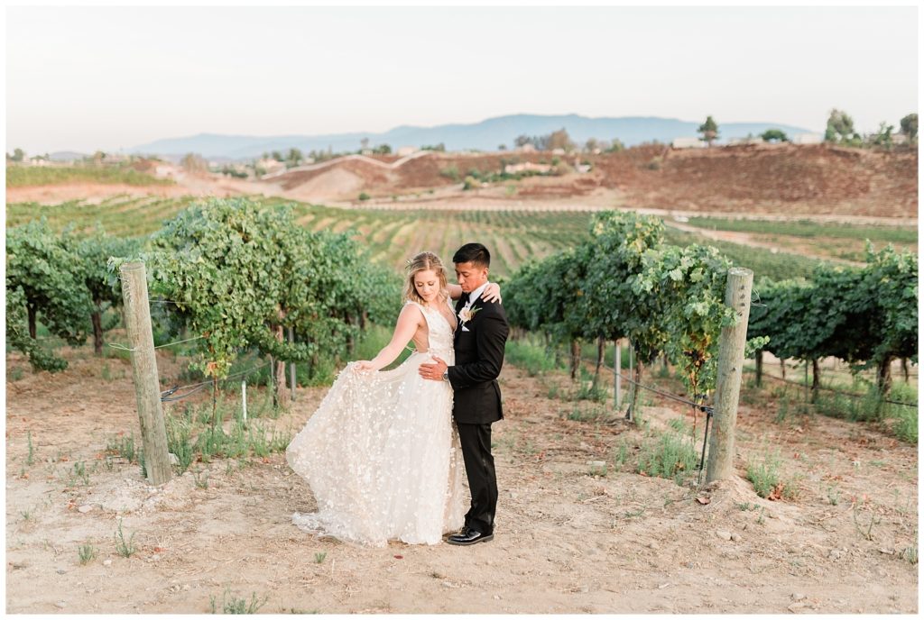 A bride and groom pose in front of the rolling vineyard hills at Avensole Winery wedding venue in Temecula, California.