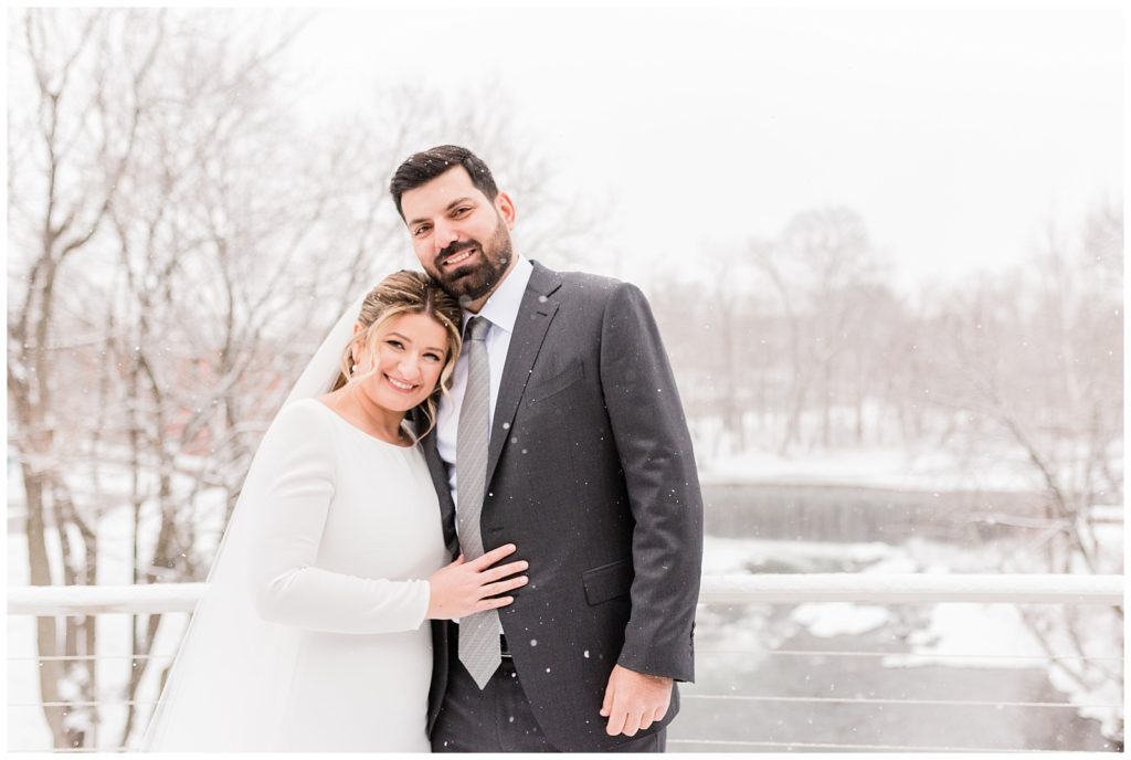 Bride and groom smiles at the camera as the snow falls around them in Beacon NY at Roundhouse Hotel winter wedding.
