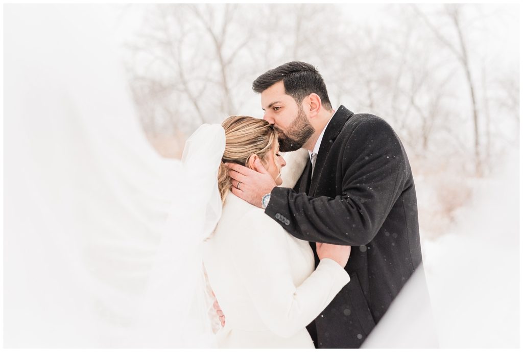A groom kisses the bride on her forehead as the snow falls around them in Beacon NY at Roundhouse Hotel winter wedding inspiration.