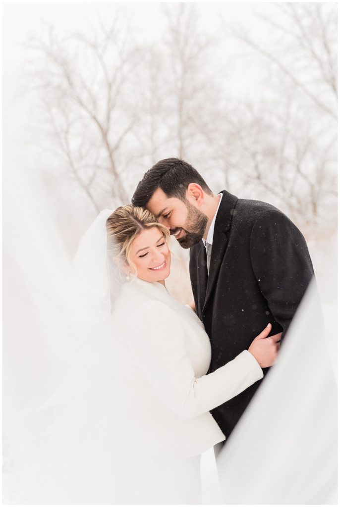 A groom nuzzles into the bride's temple with her veil sweeping in front as the snow falls around them in Beacon NY at Roundhouse Hotel winter wedding inspiration.