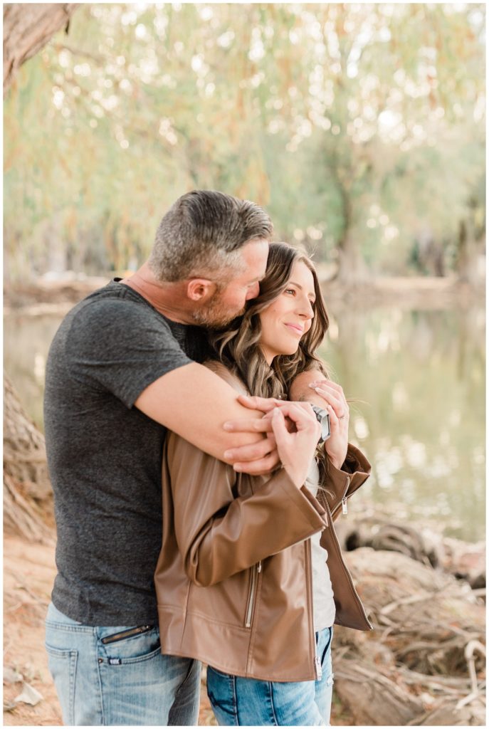 A man wraps his arms around his fiance and kisses her cheek from behind while overlooking the lake at Fairmount Park in Riverside, California.