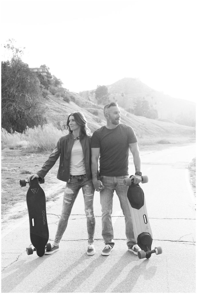 Black and white engagement photo of a couple holding hands and looking off in opposite directions while holding skateboards in their outer hands in Riverside California.