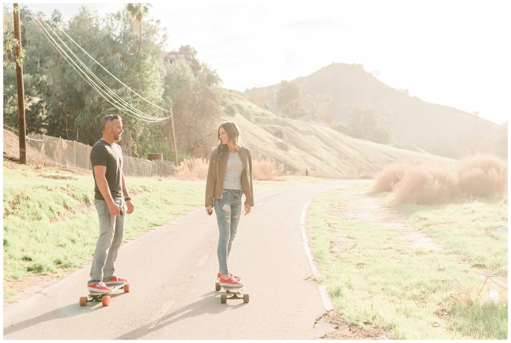A couple skateboards down a path away from a mountain while looking at each other in Riverside, California.