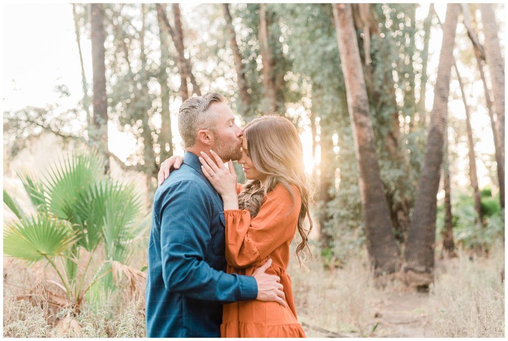 A man kisses his fiancee on the forehead while the golden sun shines behind them through the eucalyptus and palm trees in Riverside, California.