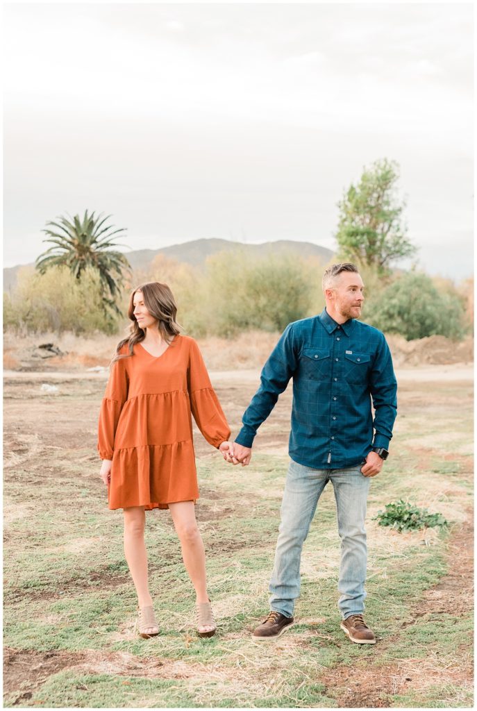 A couple holds hands looking in the opposite direction in an open field with a palm tree in the back in Fairmount Park in Riverside, California.