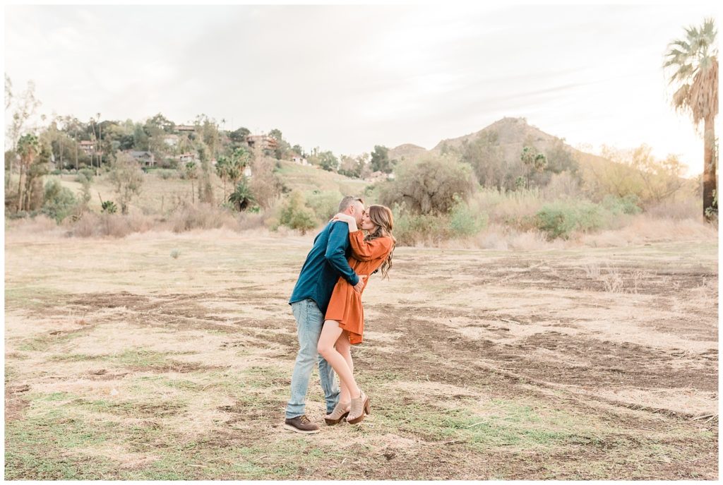 A man dips his fiancee back for a kiss in an open field at Fairmount Park in Riverside California during their engagement session.