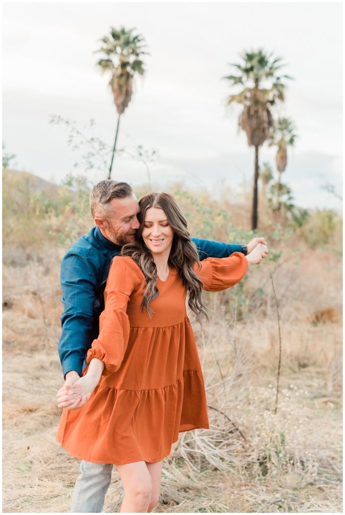 Fun adventurous couple laughs together during engagement photos holding hands with him behind her looking towards each other in Riverside California.
