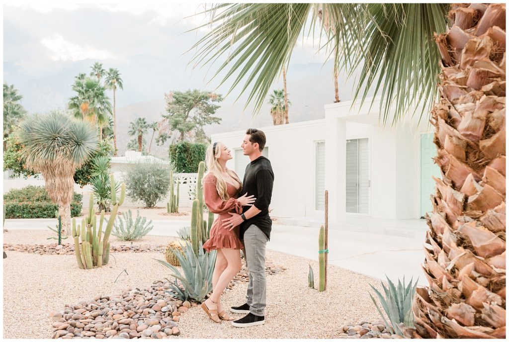 A couple stands in front of an iconic home looking at each other in Palm Springs California during their engagement session.