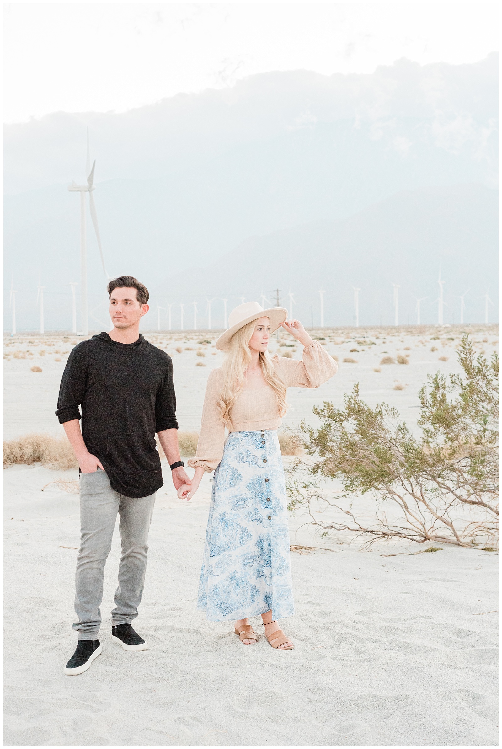 A couple holds hands in a windmill farm field in Palm Springs California during their engagement session.