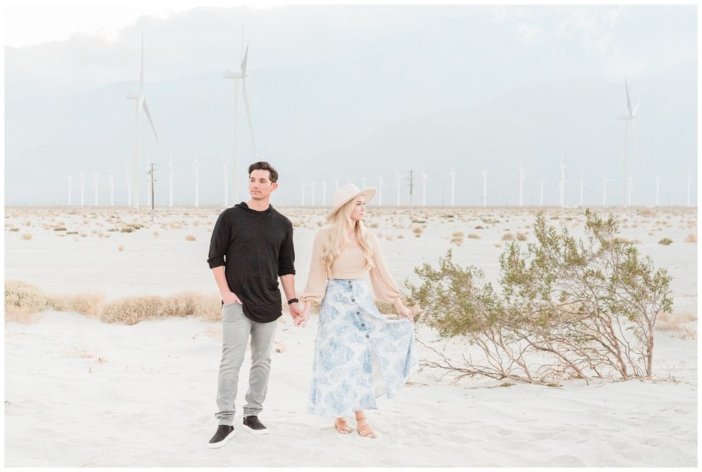 A couple holds hands looking in opposite directions in a windmill field in Palm Springs, California during their engagement session.