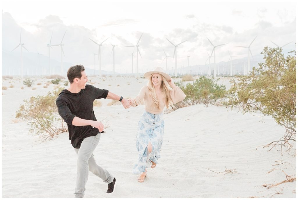 A couple holds hands running through the windmill fields in Palm Springs California during their light and airy engagement session.