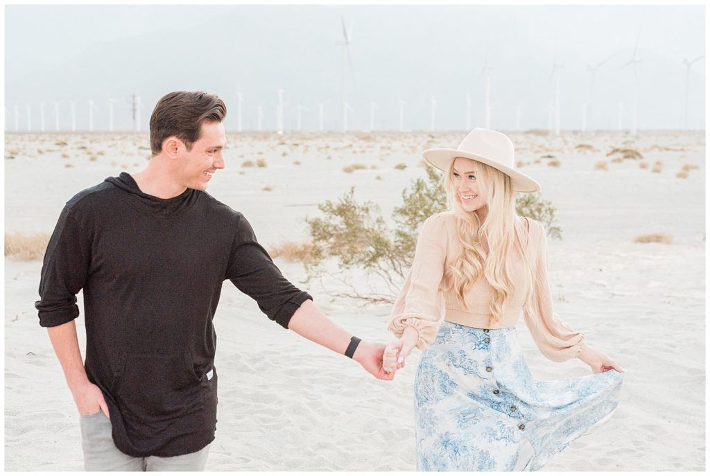 A couple smiles holding hands walking through the Palm Springs windmill fields for their engagement session.